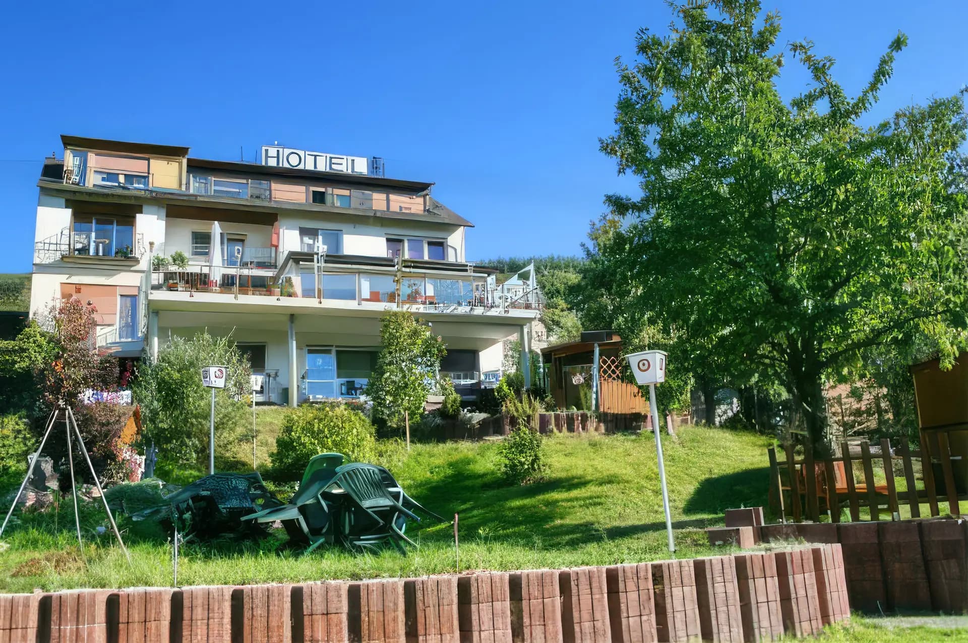 Hotel s terasami a prosklenými balkony na travnatém svahu, vpředu zahrada se složenými zelenými židlemi a velký listnatý strom