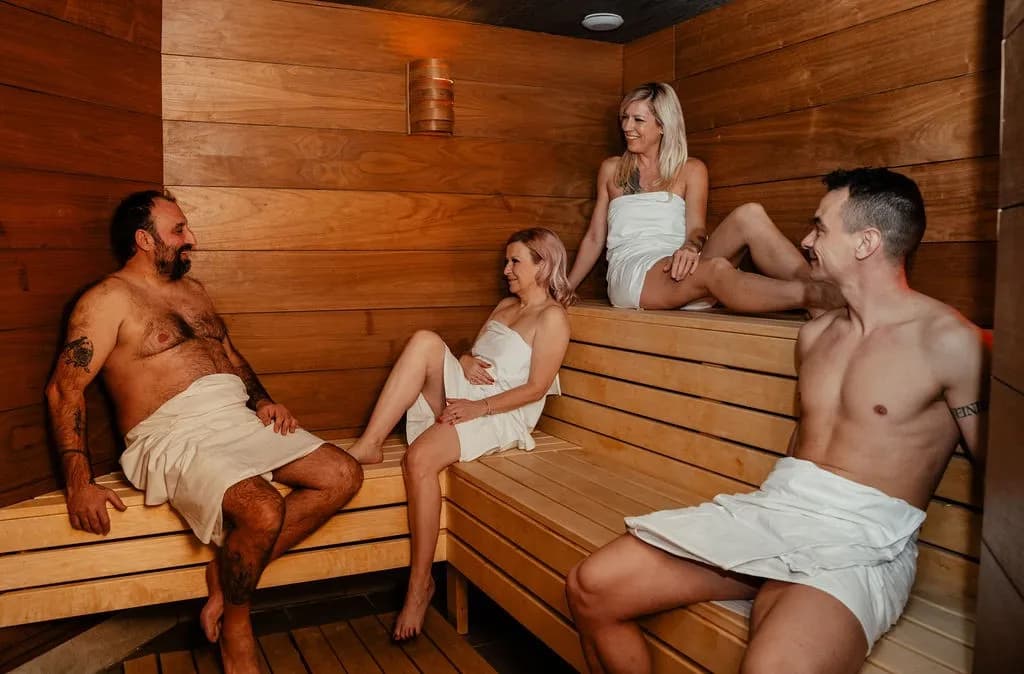 Čtyři lidé zahalení v ručnících sedí a povídají si na dřevěných lavicích v sauně