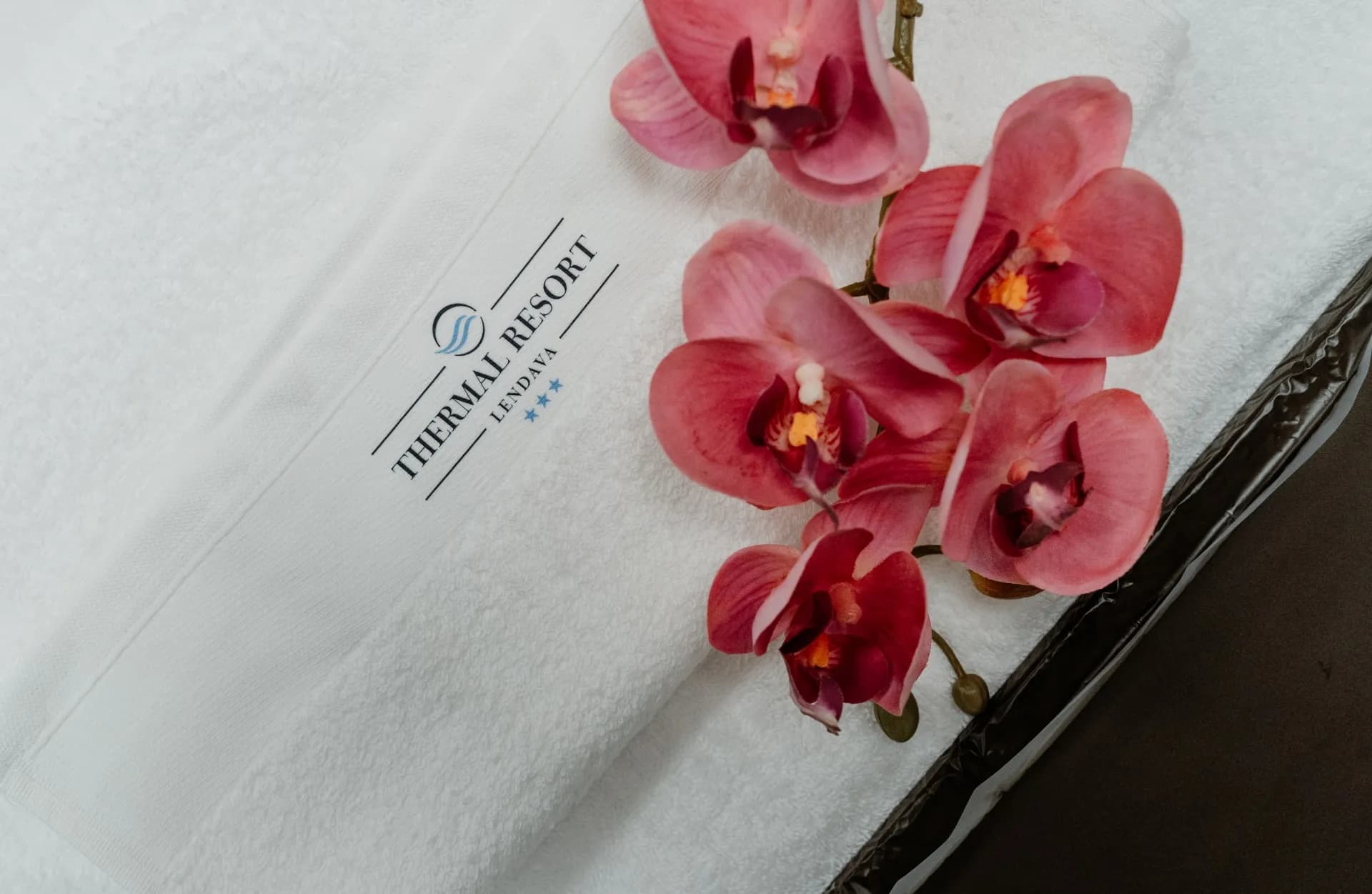 Bílý ručník s logem Thermal Resort Lendava a růžové orchideje položené na jeho okraji